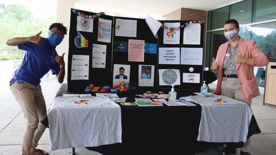 LGBTQ Affairs tabling with pamplets, signs, etc - Two advocates in masks giving the thumbs up