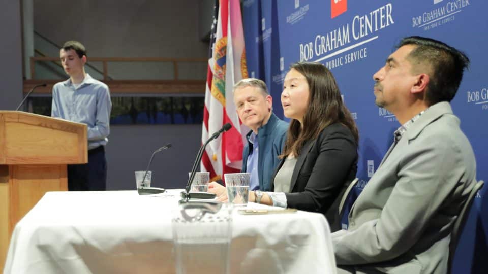 Three panelists during a talk about AI, while a young man stands at the podium in the background