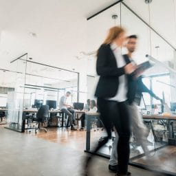 Two people walk through a modern office with glass partitions and computer stations