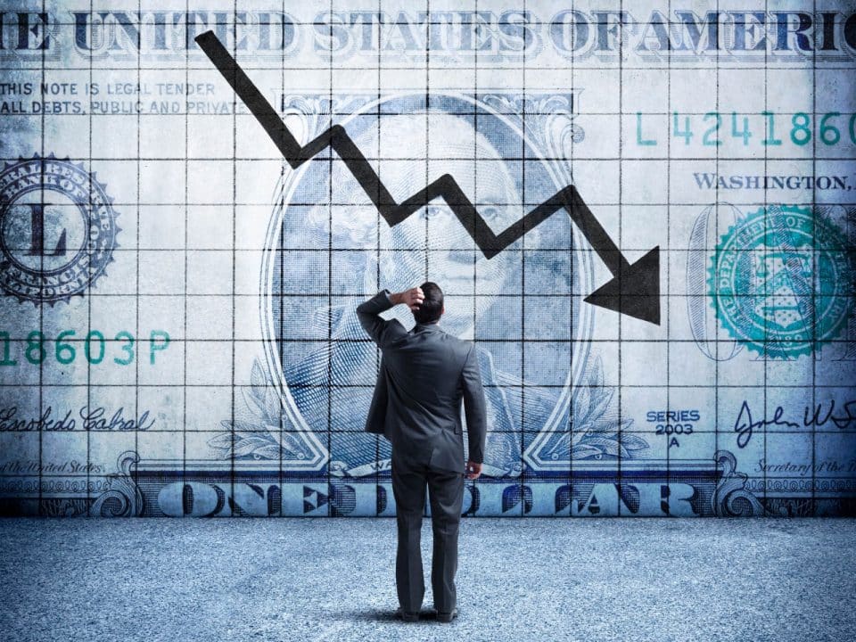 A man scratches his head while facing a giant dollar bill with a downward trending arrow on it