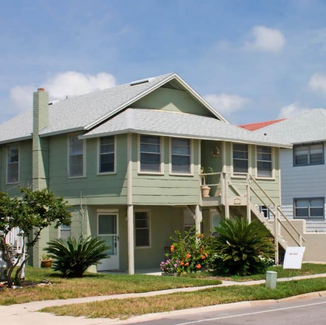 A green house with front doors on both the ground floor and second story has a For Rent sign by the street