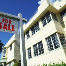 A pale yellow with light green trim art deco house in Miami with a for sale sign in front of it