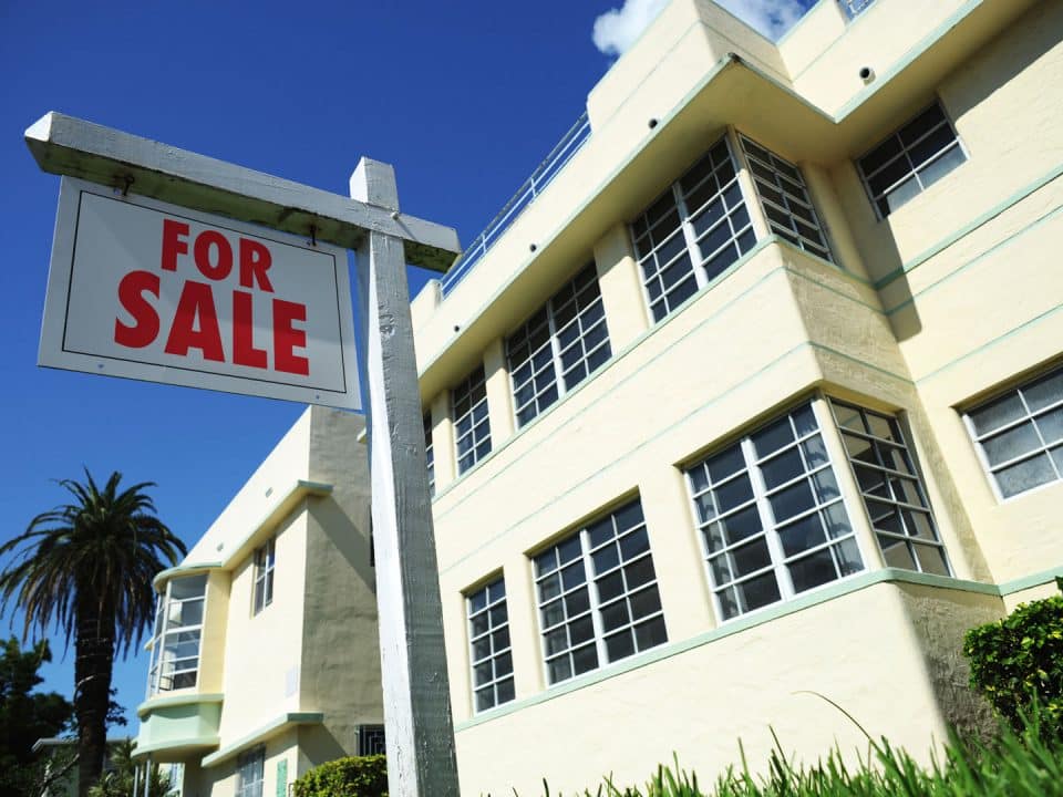 A pale yellow with light green trim art deco house in Miami with a for sale sign in front of it