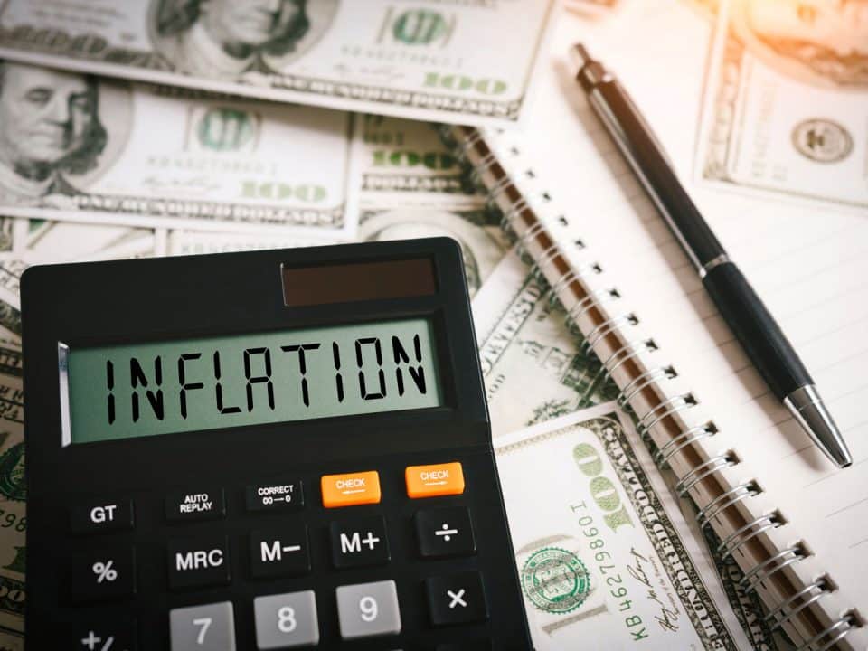 Calculator displaying 'Inflation' sitting on scattered $100 bills with a notebook and pen on the side