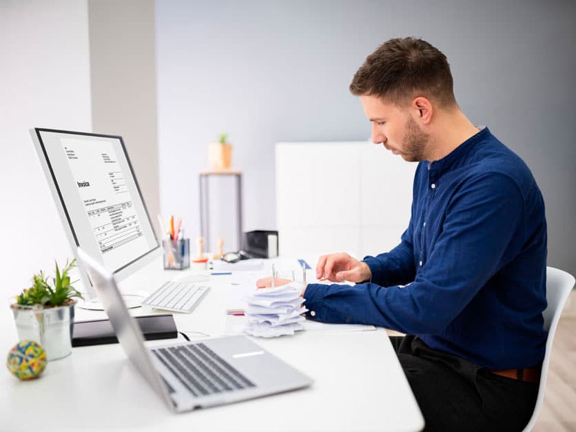 A man sits at a desk with a display, keyboard and open laptop