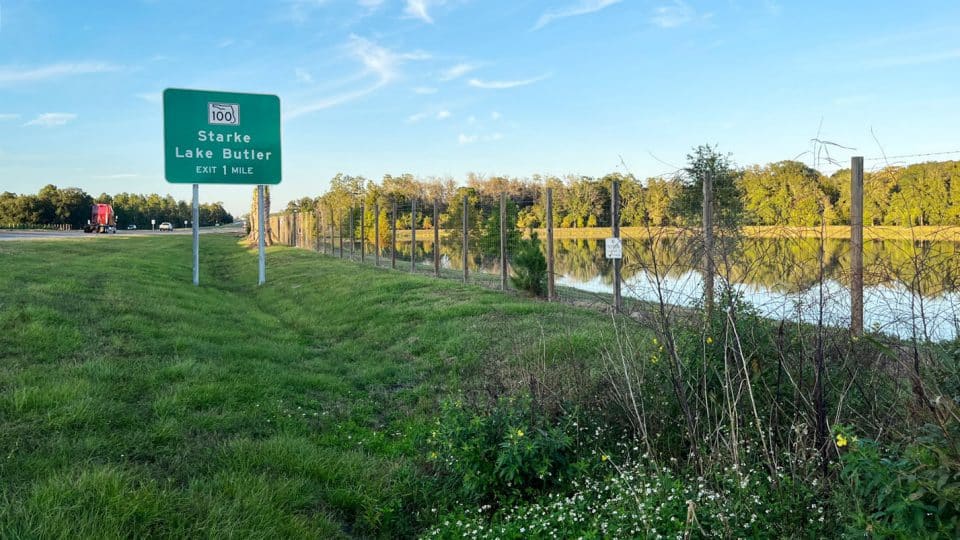 Highway sign for Starke and Lake Butler exit 1 mile, traffic on one side and water on the other behind a fence