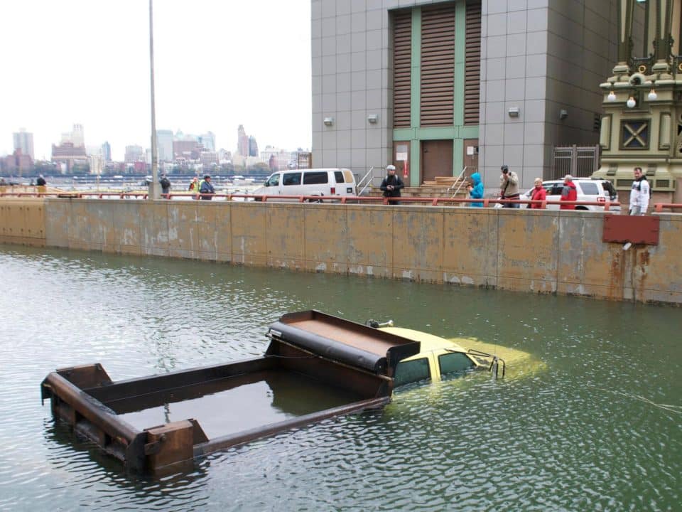 A construction truck sits submerged in water up to its windows while people look on