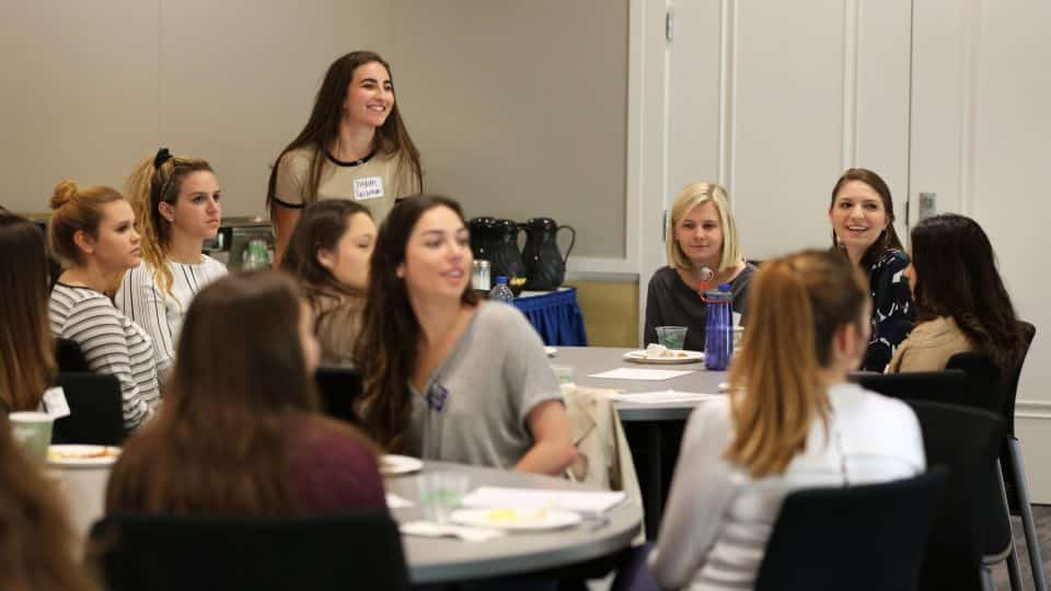 Students seated around tables listen to someone talking across the room