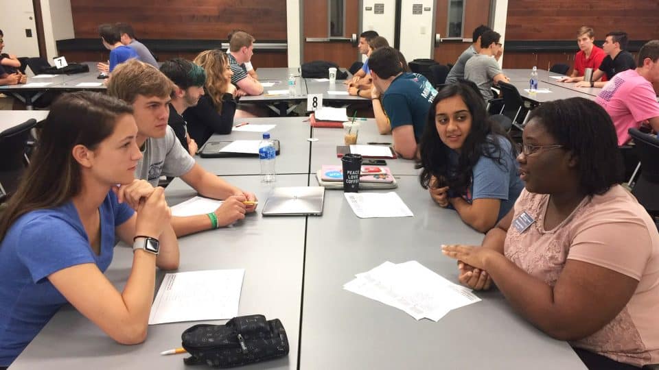 Students are grouped for discussions at tables in a classroom