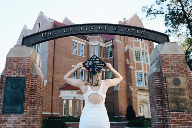 Graduate with decorated mortar board looks on at the University of Florida gateway with Heavener Hall in the background