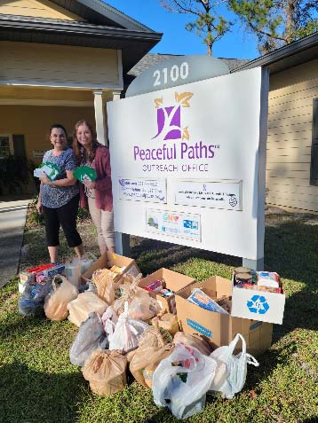 Ambassador with another individual by a Peaceful Paths sign with bags and boxes full of items stacked below it