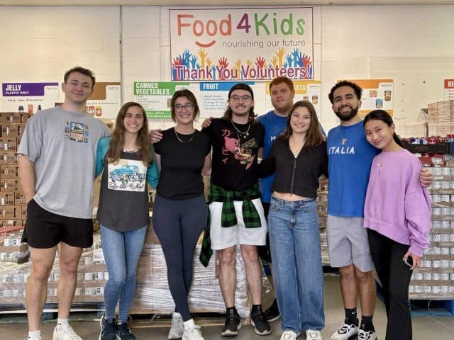 A group of Hough Graduate Ambassadors pose in front of of stacks of canned and boxed food for Food4Kids