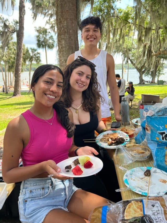 Three Hough Graduate Ambassadors enjoying food at a picnic table with Lake Wauburg in the background