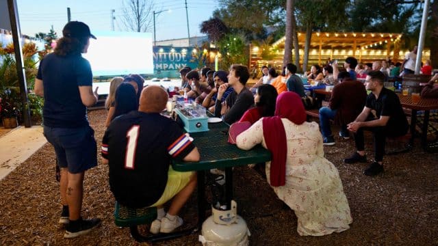 Hough Graduate Ambassadors seated at tables watch a big screen at an outdoor Super Bowl Watch Party at First Magnitude Brewery