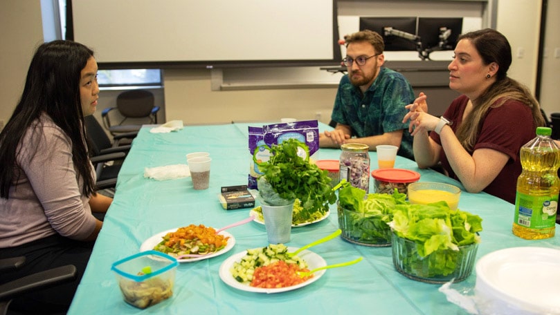 Three students sit at a table with plates and ingredients for arepas