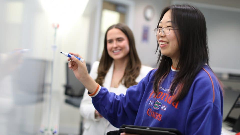 A student holds a blue marker up to a dry erase board as another student looks on