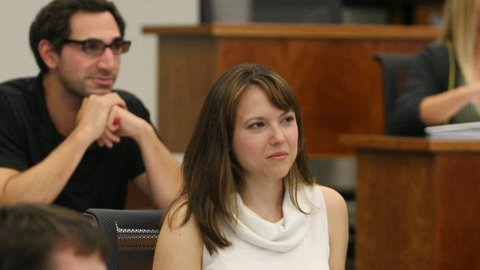 A woman in a classroom listens to the instructor as a man in the background also listens