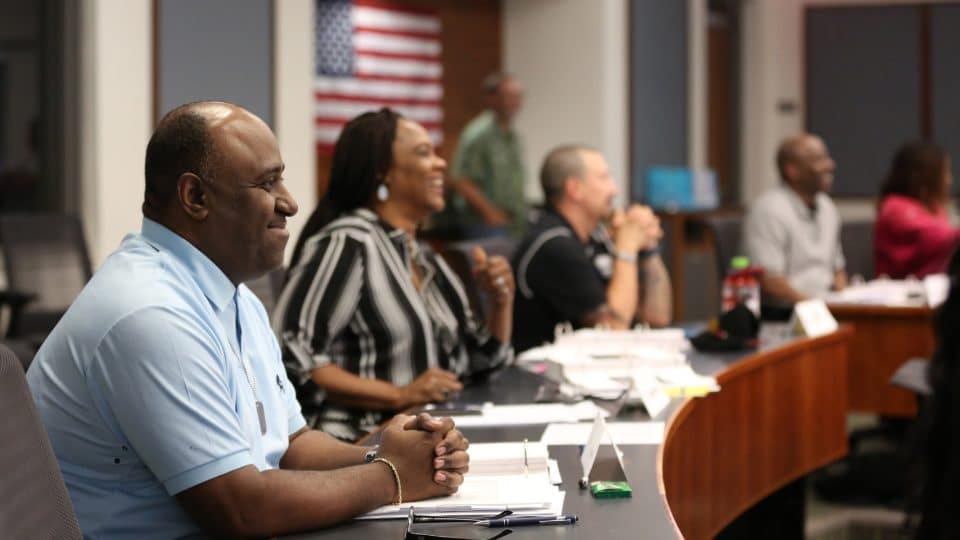 A man smiles in a classroom as other students in the background do the same