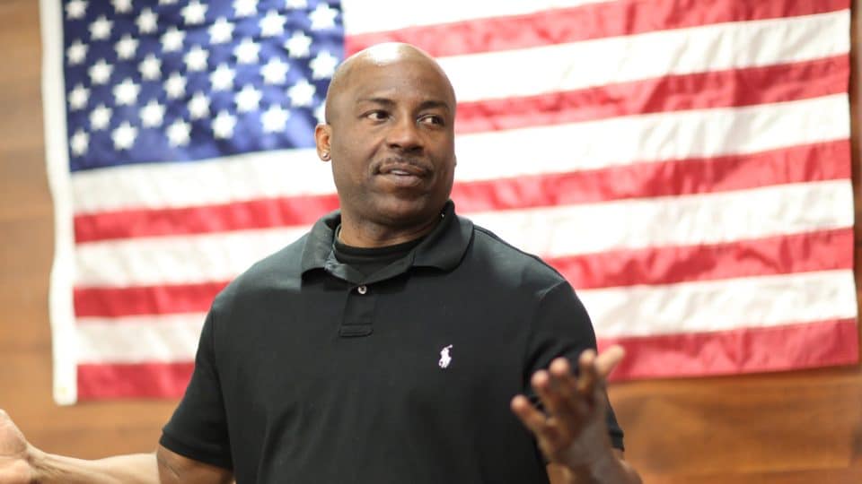 A man addresses the classroom with a U.S. flag on the wall behind him