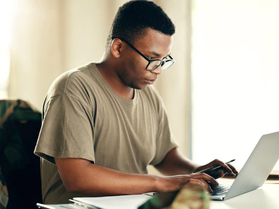 A man works on a laptop with a military jacket hung on his chair