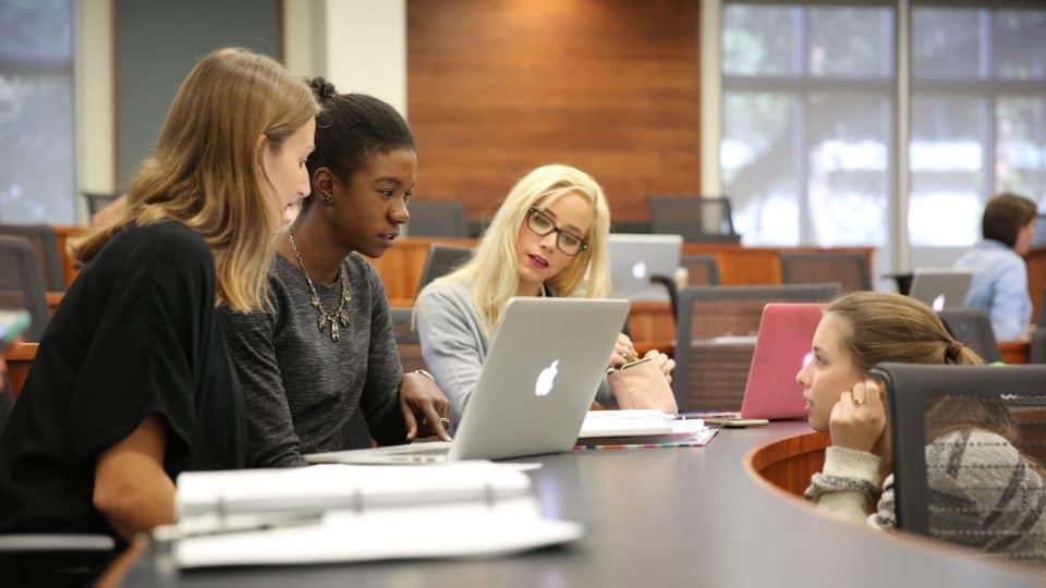 Four students in a classroom having a discussion with a laptop and notebooks open on the curved table