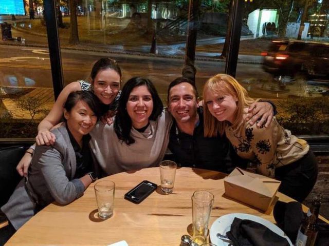 Five people smile for the picture while gathered around a table in the evening with a street outside the window behind them