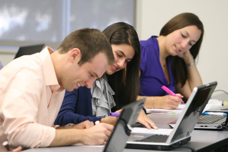 Three students write on papers at a table with their laptops open in front of them