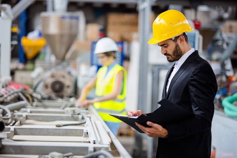 Man in a suit and yellow hard hat looking at parts in a factory and taking notes