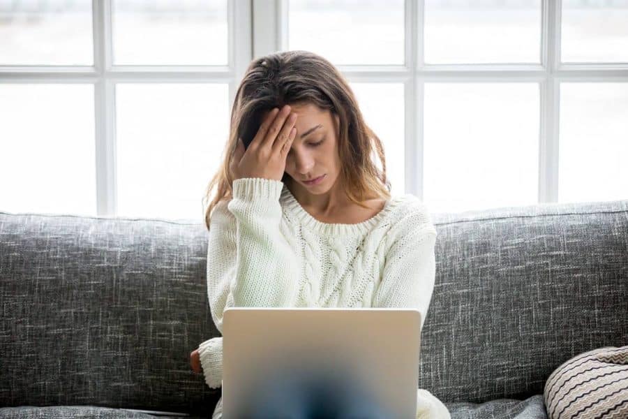 Young woman sitting on a couch looking at a computer. She holds her hand up to her forehead looking stressed.