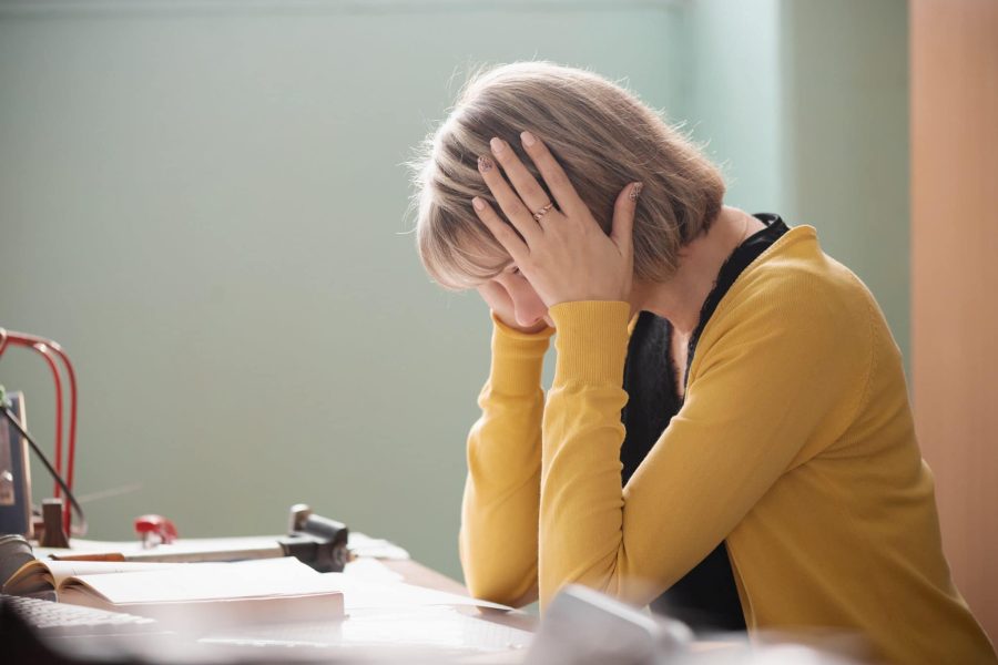 Overworked teacher is sitting at her desk with her hands on the sides of her head as she looks at papers