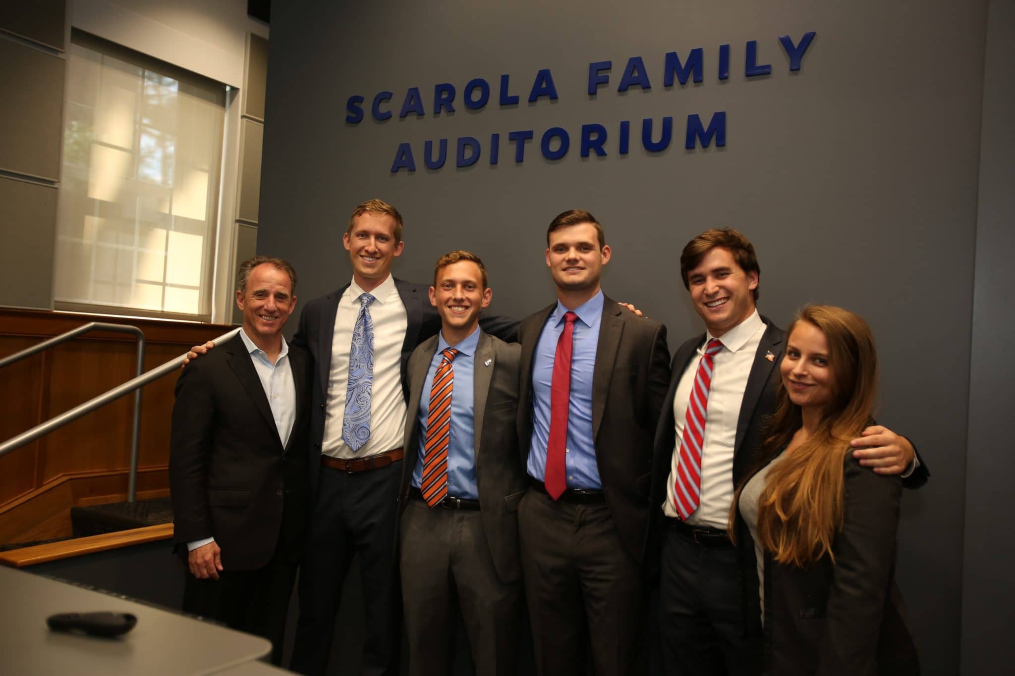 Rick Scarola with five students posing for a photo in front of the Scarola Family Auditorium sign