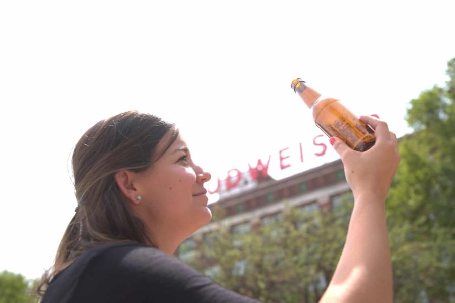 Lexie Cegelski holds a beer bottle high as if examining it with a Budweiser sign on a building in the background