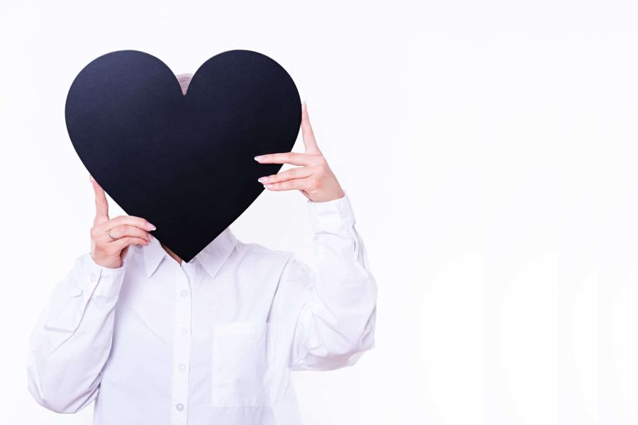 Woman in a white button-up shirt on a white background holding black cutout of a heart in front of her face