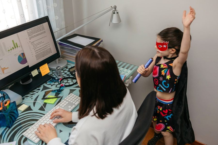 A woman at a computer and working from home with her daughter singing and playing by her side.