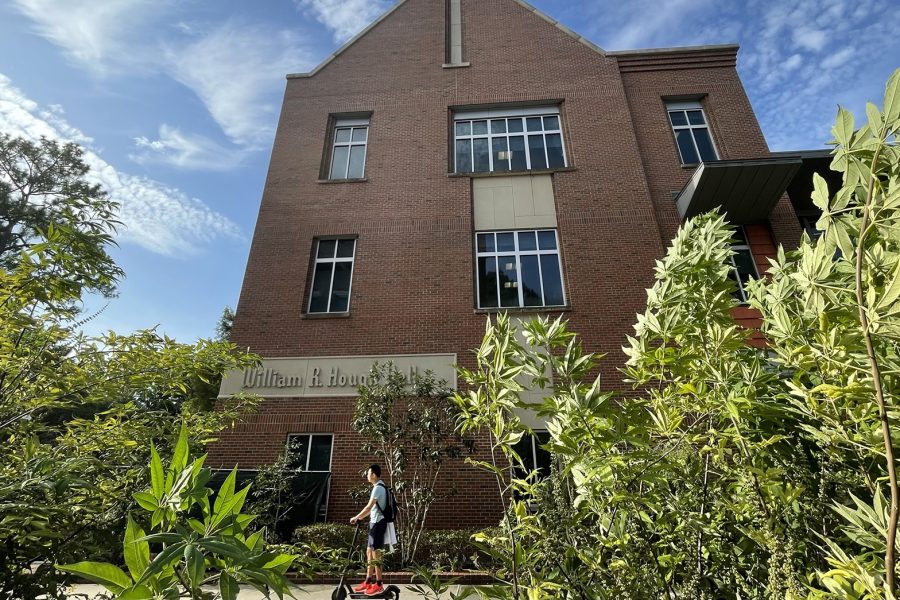 Hough Hall surrounded by greenery on a sunny day as a student on a stand-up scooter rolls by