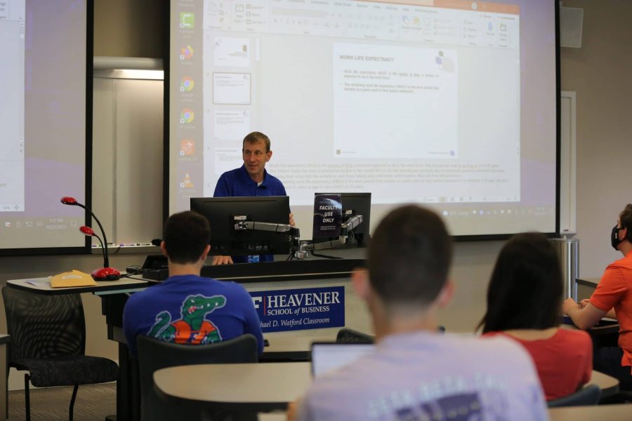 John Banko stands at the front of a classroom in Heavener Hall. Students can be seen from behind watching.