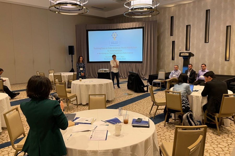 Conference room with a large presentation screen and several round tables. A group of people ask questions to two presenters of a research presentation.