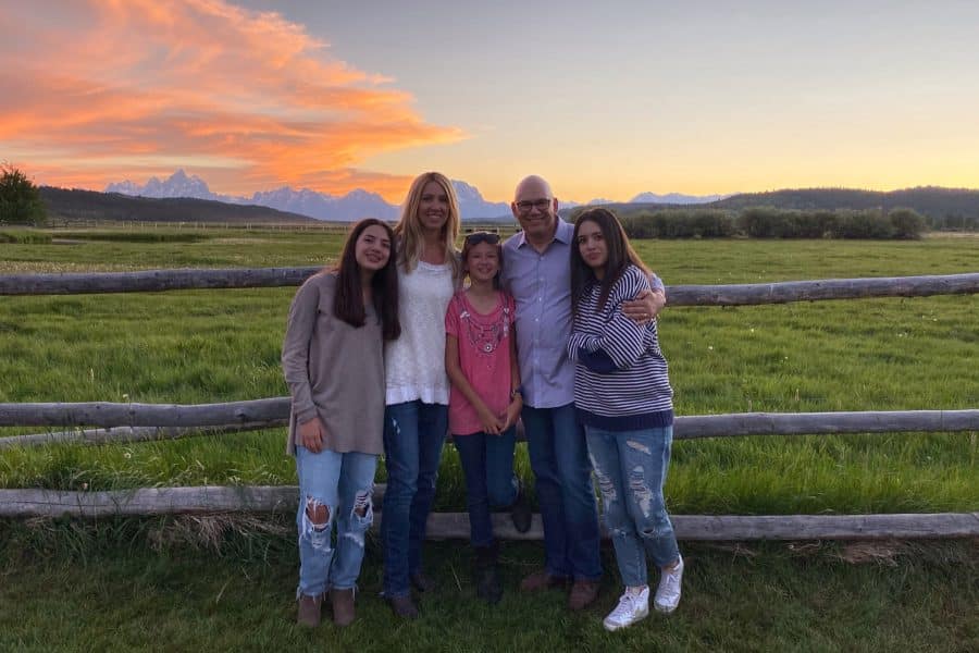 A family of five, mother, father and three young daughters, pose for a photo by a wooden fence at sunset with mountains in the far background