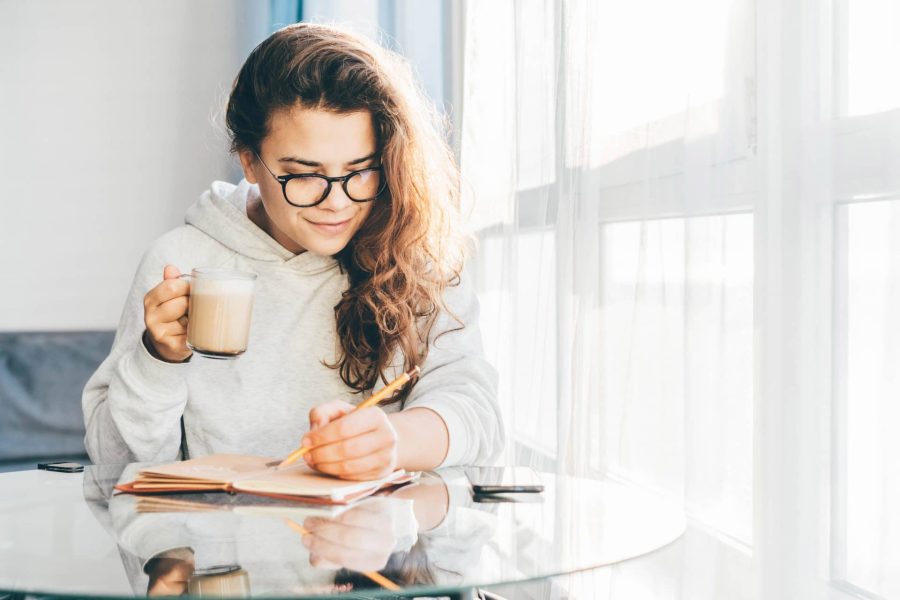Woman sitting at a table with cup of coffee and writing notes in a paper notebook.