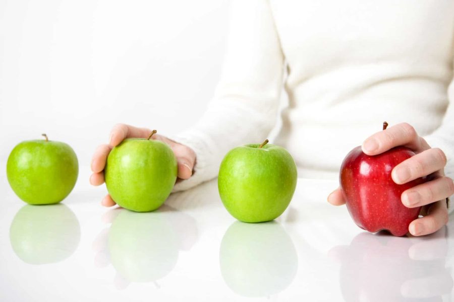 Four apples, three green and one red, sit in line with a person's hands on one green and one red apple as if trying to make a selection between the two.