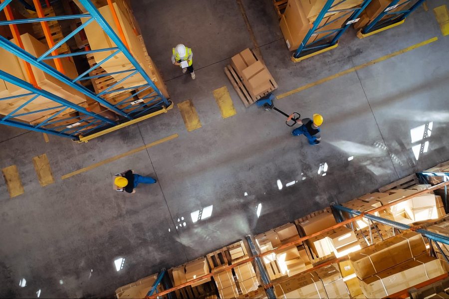 Top-down view: In warehouse people working, pallet jack operator pulls pallet with cardboard boxes on it.