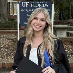 Caroline Murphy in graduation attire holds her cap in front of the Warrington College of Business sign