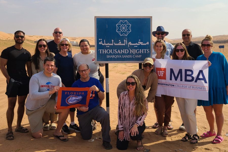 A group of about 15 people, including students and alumni, pose for a photo in the Omani desert while holding a UF MBA and University of Florida flag.