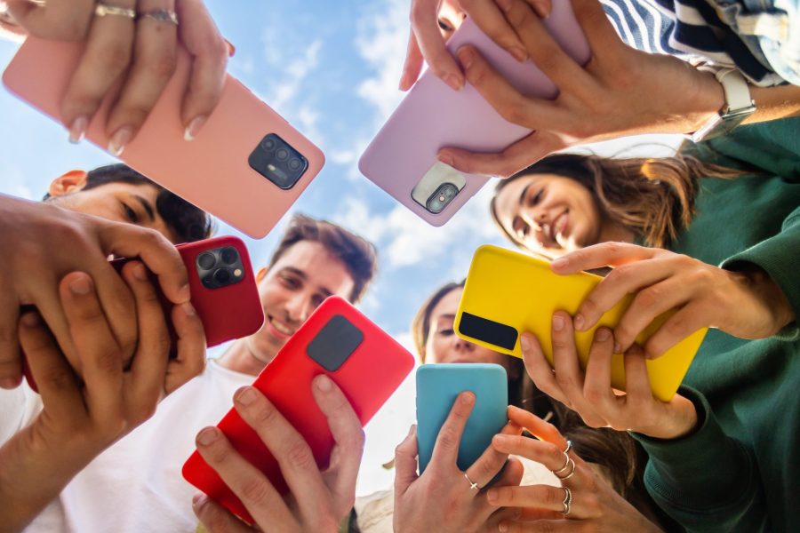 Young group of people using mobile phone device standing together in circle outdoors. Millennial friends addicted to social media app, betting or playing video game on platform online.