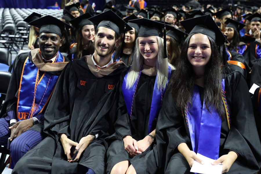 Four students, two male and two female, sit together in graduation regalia during a graduation ceremony.