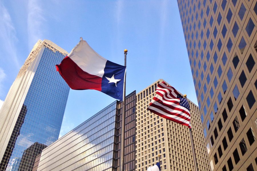 Low angle view of United States of America flag and Texas state flag with tall buildings behind them.