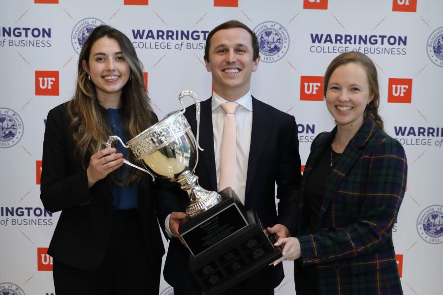 Three students wearing business attire hold a large trophy cup