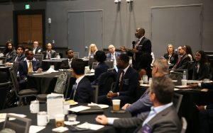 A man in a suit stands up in a large group watching a presentation while asking a question with a microphone.