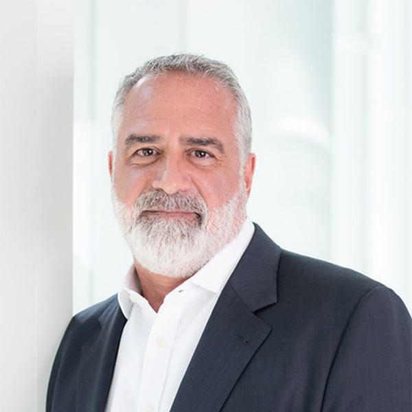 Kenny Polcari, wearing a white dress shirt and dark blazer, stands in front of a softly lit modern background with a confident expression.