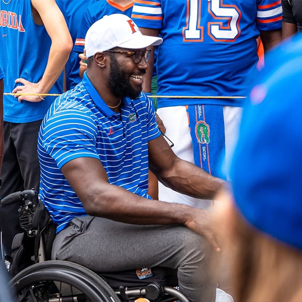 Patric Young, wearing a University of Florida polo and white cap, smiles while seated in a wheelchair during a public event surrounded by Gators fans.
